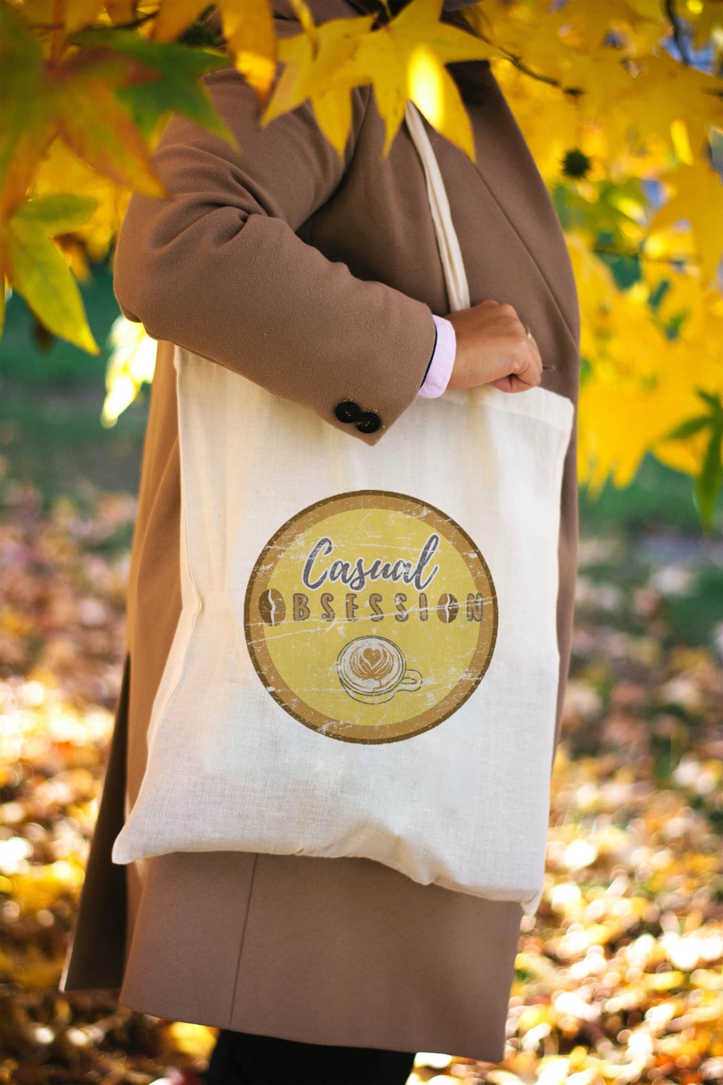 A wooded scene and lady holding a natural colour canvas tote bag with a unique, retro graphic display showing a coffee cup and the words "Casual Obsession"